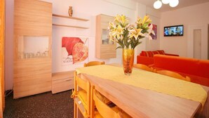 Et spiseområde med et træbord, en gul dug og en vase med blomster. Der er orange stole og et fjernsyn på væggen.