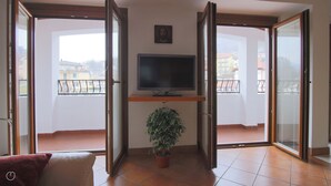 A living room with a TV on a stand, a potted plant, and a balcony with a view of buildings. - Italianway - Piazza Nuova 3 (Centro Valle Intelvi)
