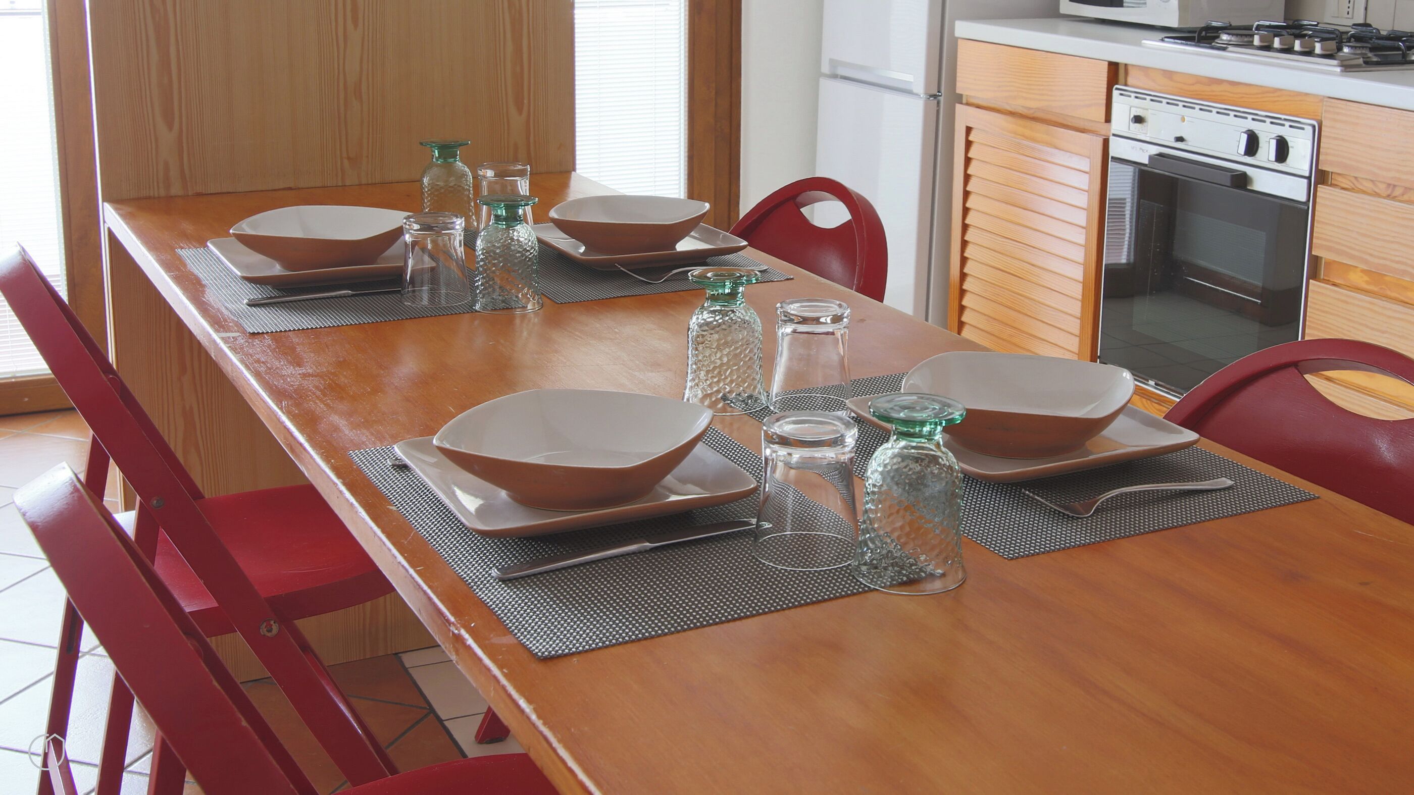 A dining table set with bowls, plates, and glassware, surrounded by red chairs in a kitchen with wooden cabinets and a stove.