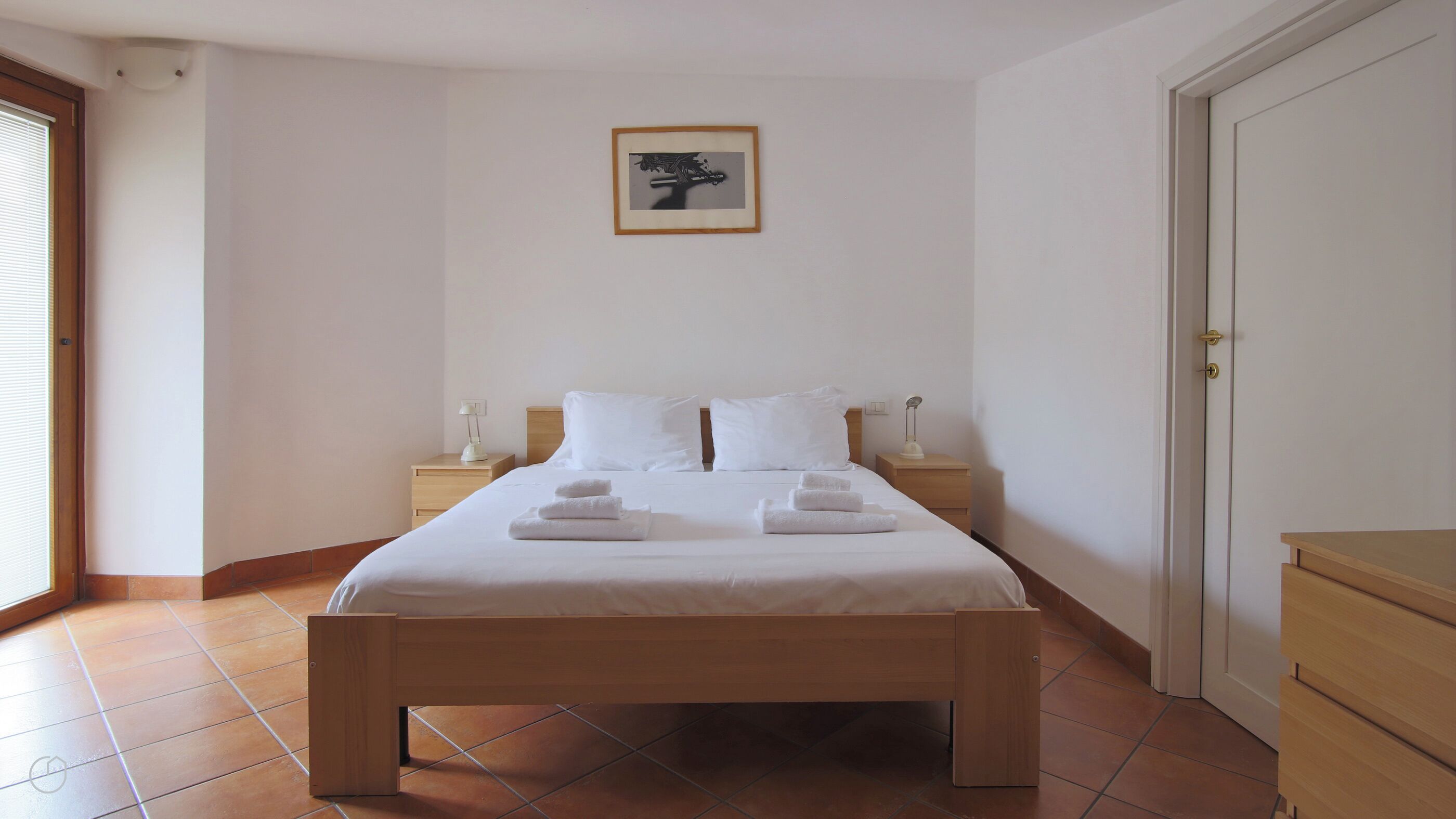 A neatly made bed with white linens and towels, a bedside table with a lamp, and a framed picture on the wall.