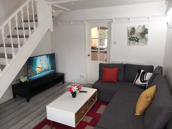 A living room with a staircase, a flat-screen TV, a grey sofa, a white coffee table, and a red and beige rug.