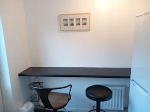 A small kitchenette with a black countertop, a chair, a radiator, and a framed picture on the wall.