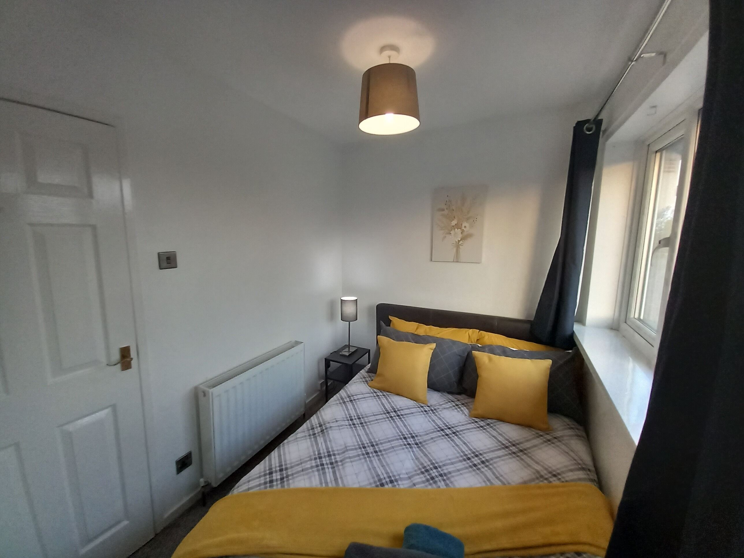 Un dormitorio con una cama, una mesita de noche, una lámpara, un radiador, una ventana con cortinas y una puerta.
