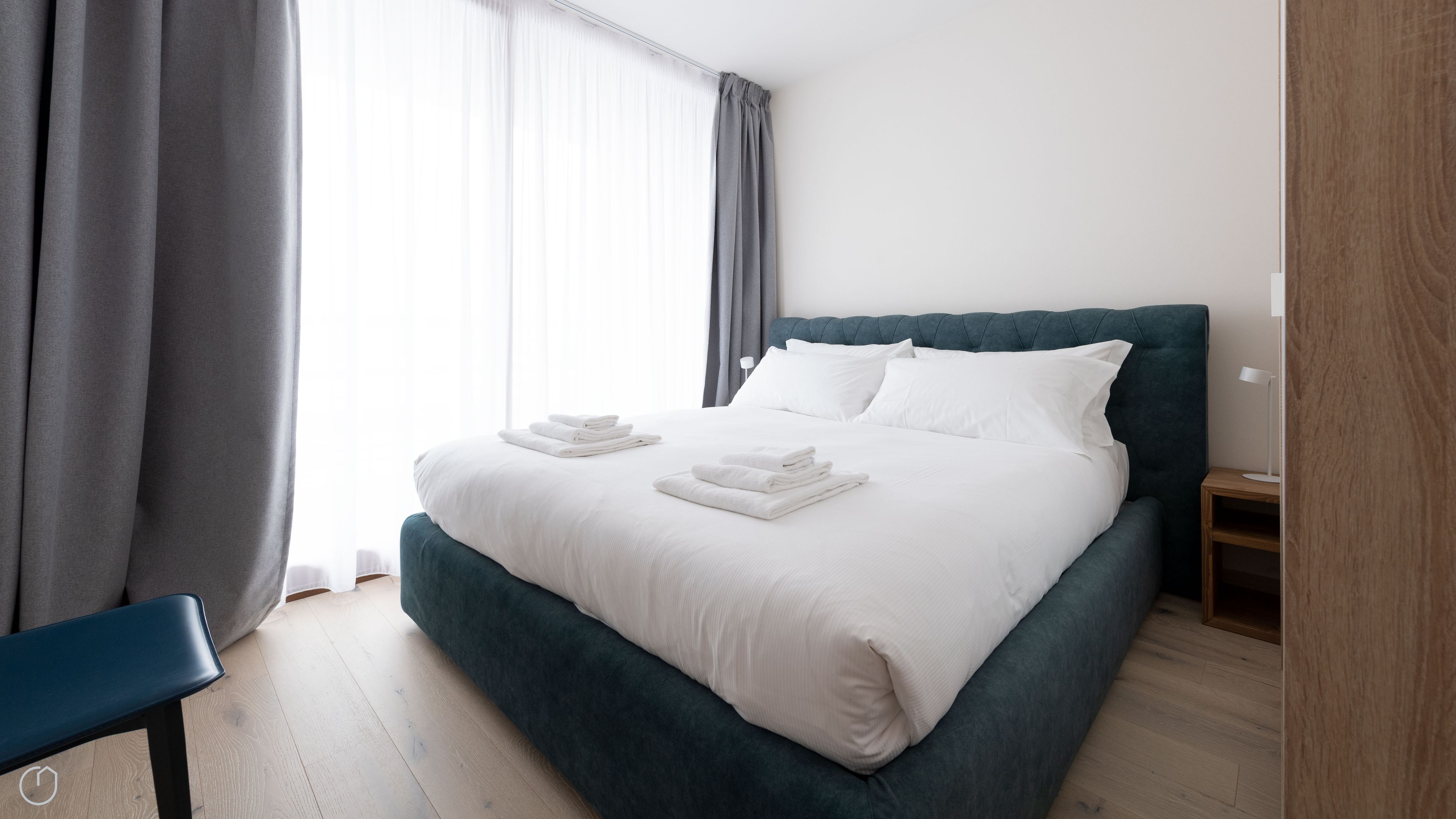 A neatly made bed with white linens and towels, a dark green upholstered headboard, and a wooden nightstand with a lamp.