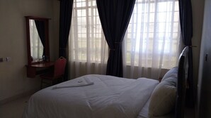 Ett hotellrum med en säng, ett skrivbord, en stol, en spegel och stora fönster med gardiner.