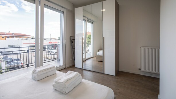 Un dormitorio moderno con un amplio ventanal corredera que da a un balcón, una cama con toallas blancas ordenadas y vista a un edificio comercial.