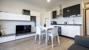 Una cocina moderna con armarios blancos, horno empotrado, microondas y cafetera. Hay una mesa de comedor con sillas blancas y un jarrón con flores.