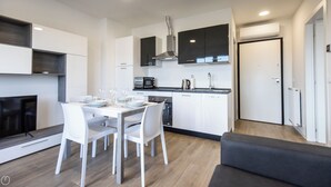 Una cocina moderna con armarios blancos, encimera negra y microondas integrado. Hay una mesa de comedor con sillas y un televisor en una esquina.