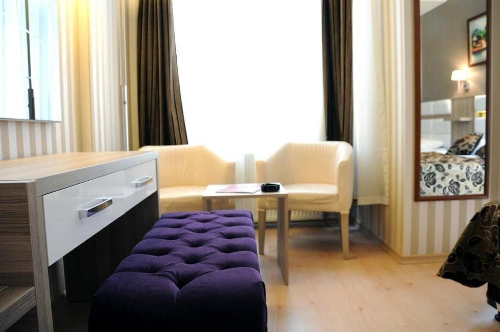 Habitación de hotel con un escritorio de madera, dos sillas, un reposapiés morado, una mesita pequeña y un espejo con motivos florales.