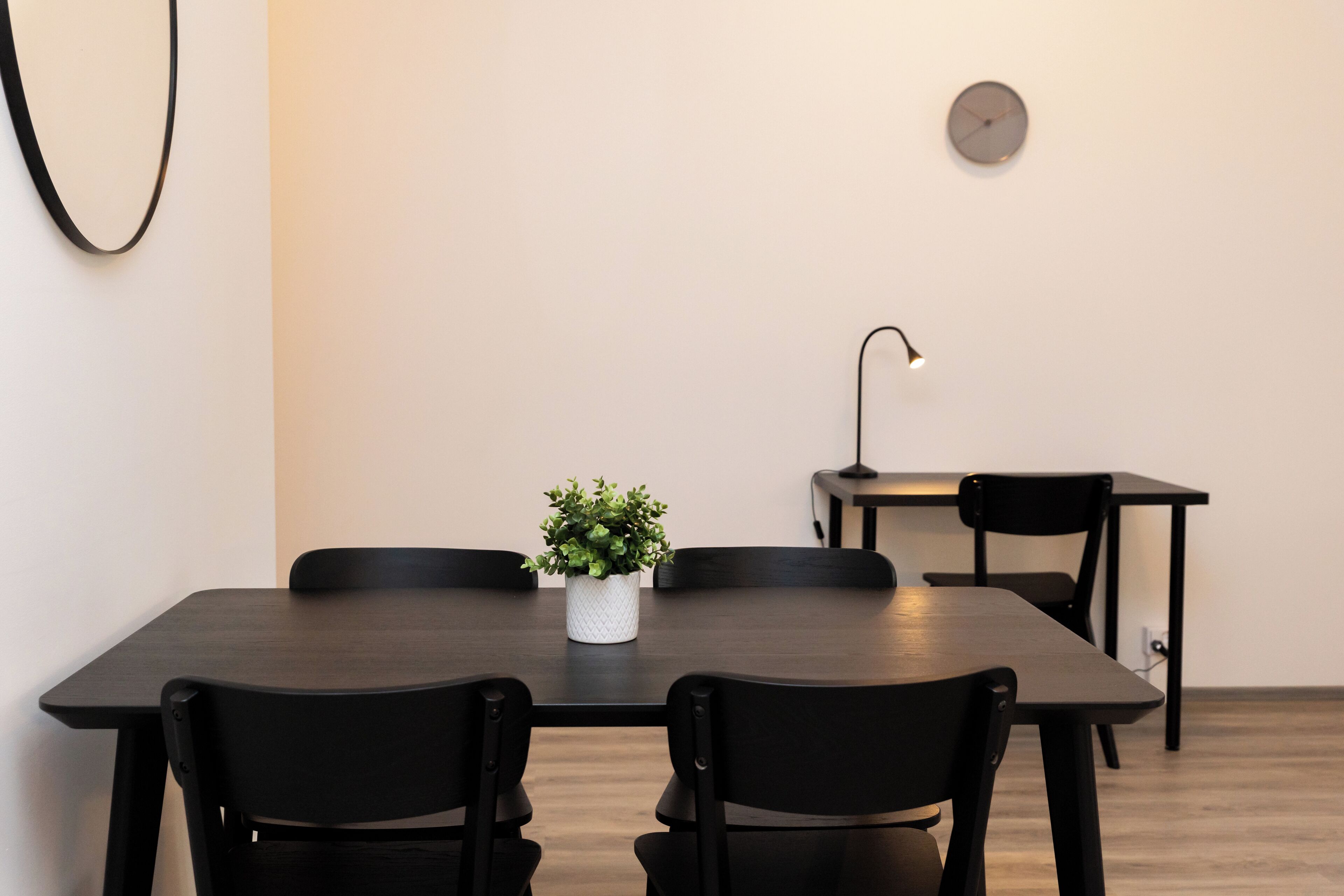 Un comedor moderno con una mesa de madera oscura, sillas negras, una pequeña planta en maceta, un reloj de pared y un escritorio con una lámpara.