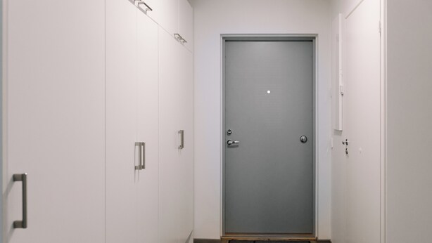 Un pasillo estrecho con una puerta gris al final, paredes blancas y suelo de madera.