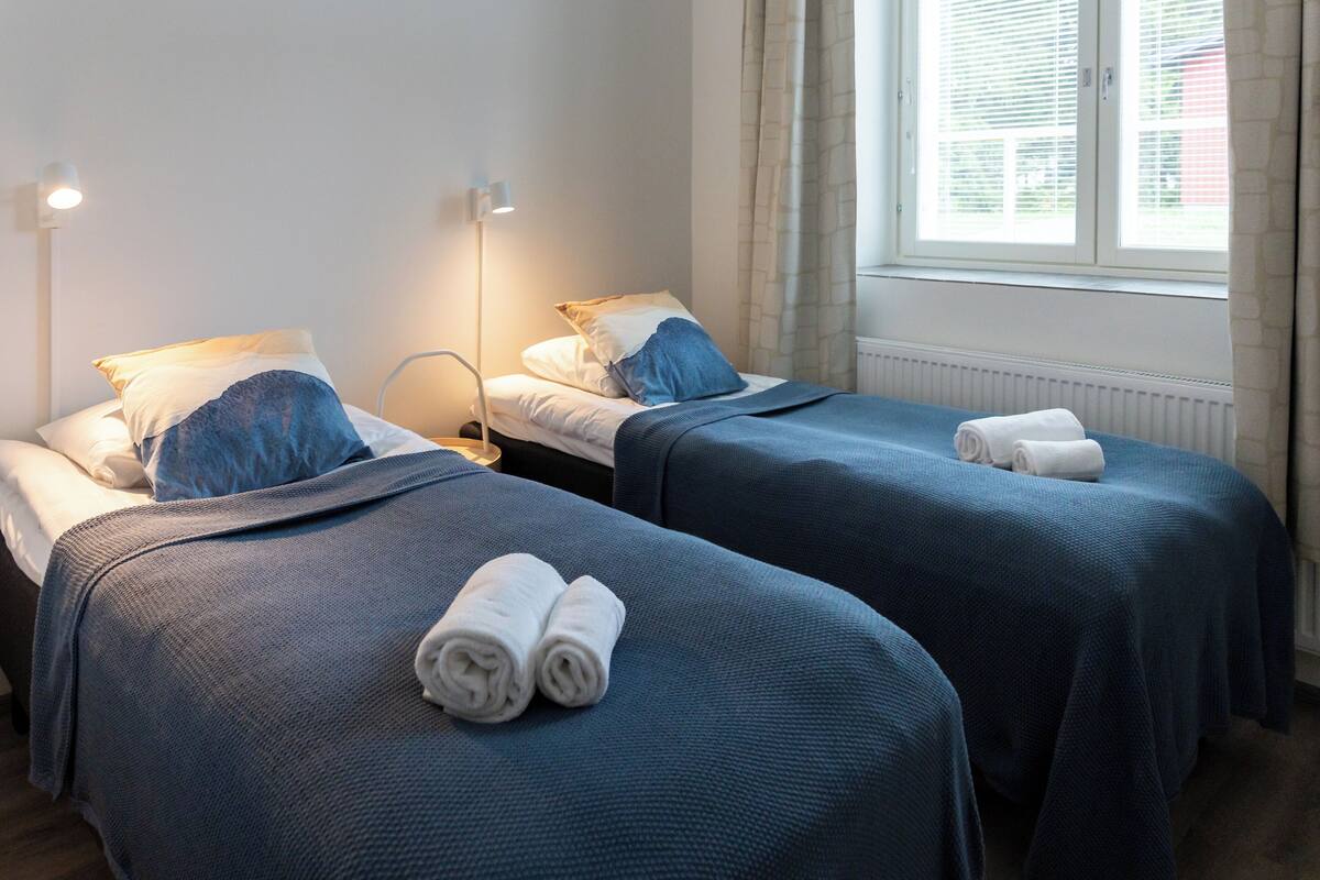 Ein Hotelzimmer mit zwei Betten, jeweils mit blauer Bettdecke und Kissen, sowie einem Fenster mit Vorhängen.