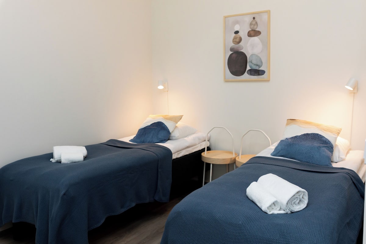 Ein Hotelzimmer mit zwei Einzelbetten, jeweils mit blauer Bettdecke und weißen Kissen, einem kleinen runden Tisch und einem an der Wand hängenden Kunstwerk.