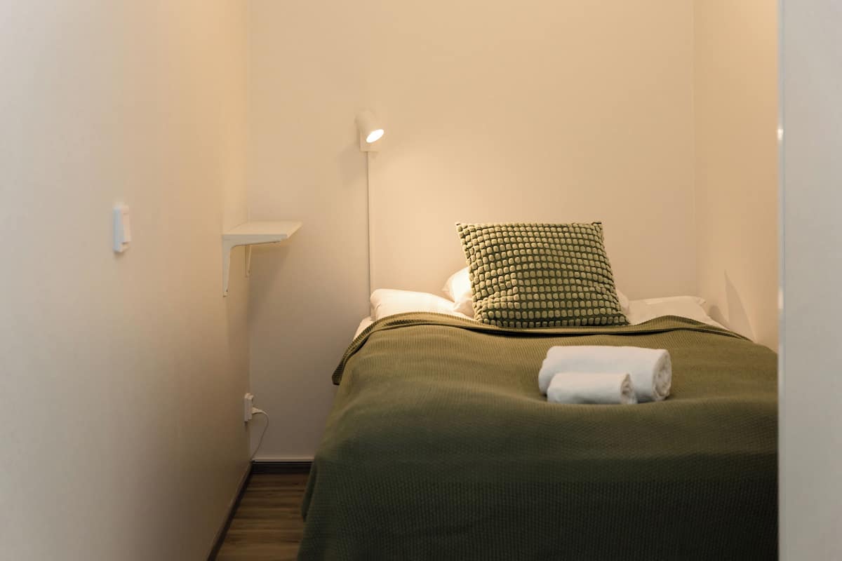 Ein kleines, ordentlich bezogenes Bett mit grüner Decke und einem gemusterten Kopfkissen, eine Nachttischlampe und eine weiße Steckdose an der Wand.