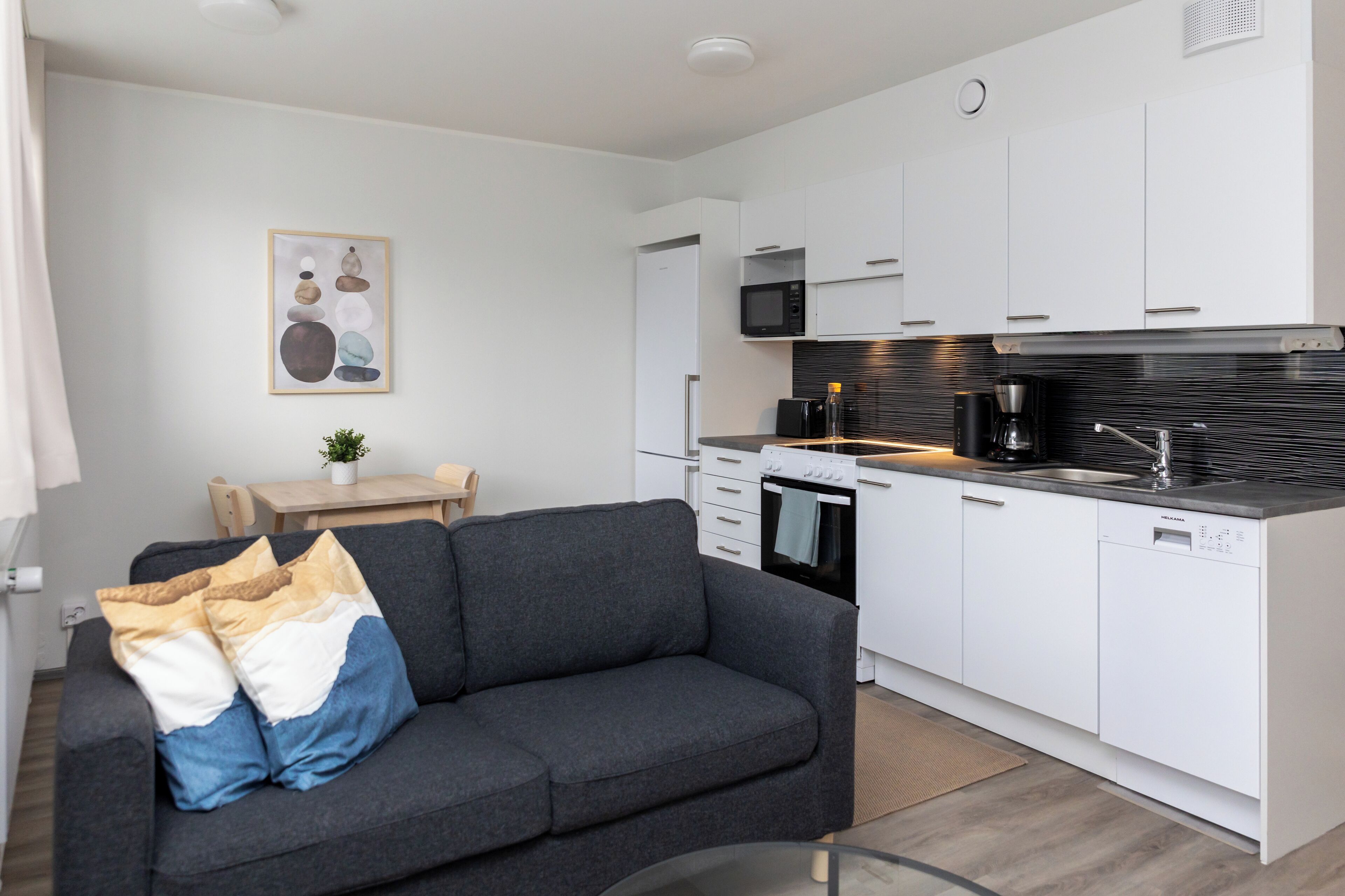 Eine moderne Küche mit weißen Schränken, einer Mikrowelle und einer Kaffeemaschine. Im Wohnbereich befindet sich ein graues Sofa mit Kissen.