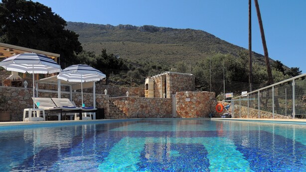 A swimming pool with clear blue water, surrounded by lounge chairs and umbrellas, with a stone wall and a mountain in the background.
