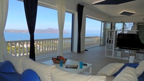 A modern living room with a sofa, coffee table, and a flat-screen TV. A balcony with a railing and a view of the sea is visible.