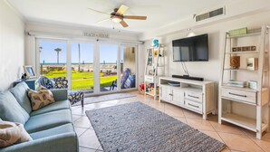 A living room with a sofa, a television, a bookshelf, and a view of the beach through the sliding glass door. - Coquina Condos: Ocean View, Ground Floor (St. Augustine)