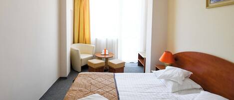 A hotel room with a bed, a chair, a small table, a lamp, and a painting on the wall.