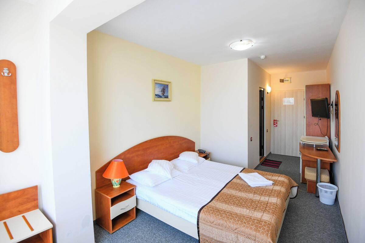 A hotel room with a bed, a nightstand, a lamp, a desk, a chair, a mirror, and a small window.
