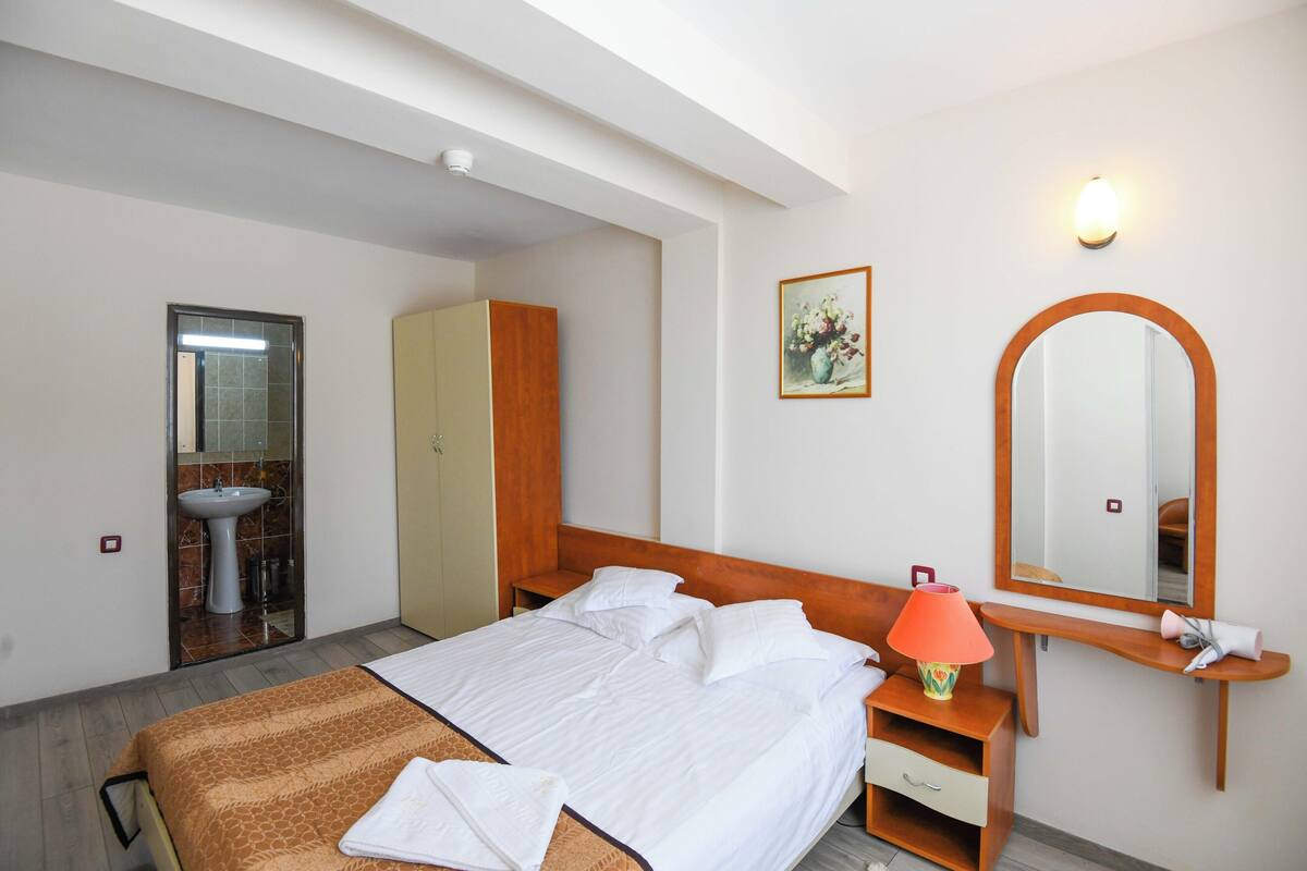 A hotel room with a bed, a bedside table with a lamp, a mirror, a wardrobe, and a bathroom with a sink and mirror.