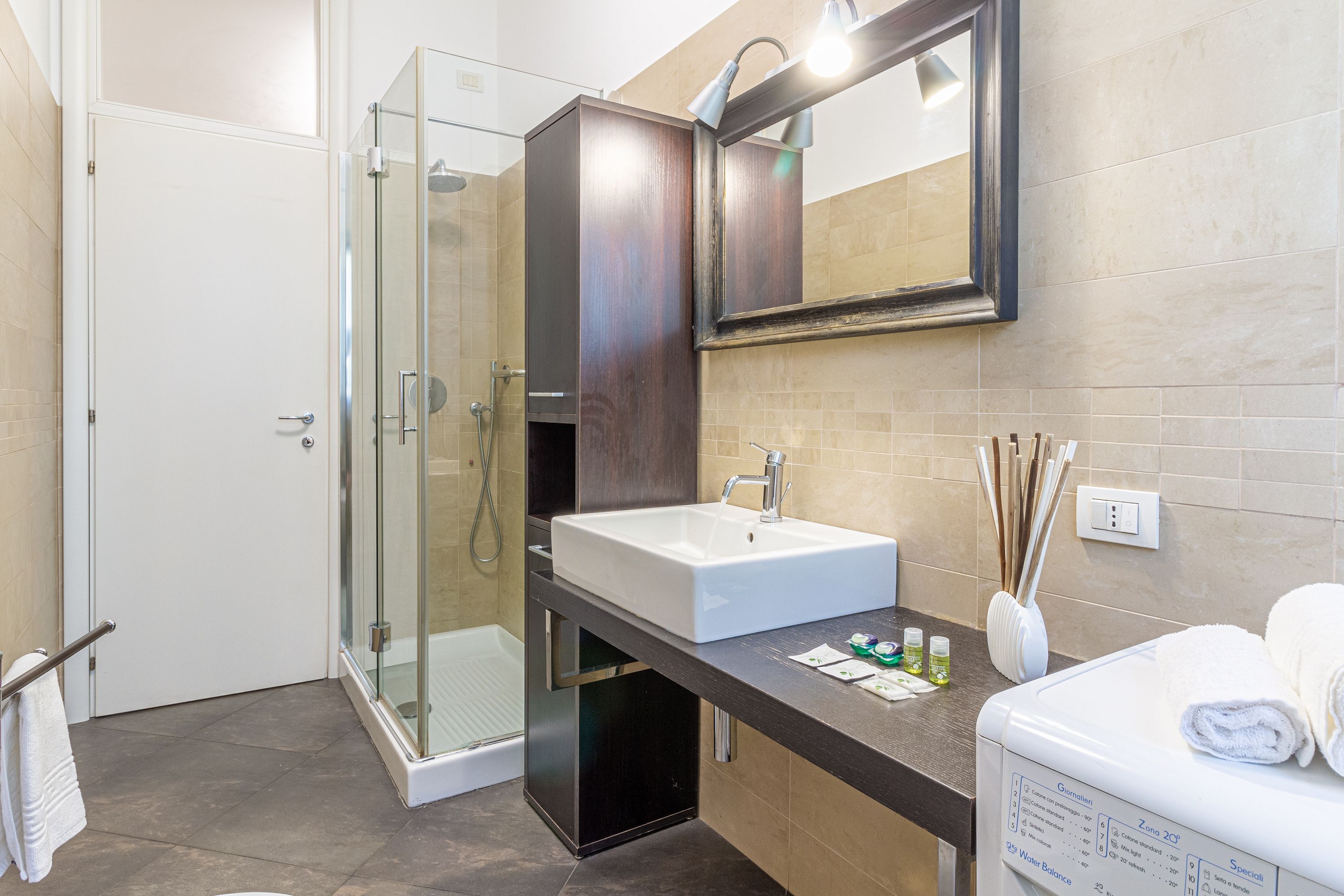 Un bagno moderno con cabina doccia in vetro, lavabo bianco e mobile contenitore in legno scuro.