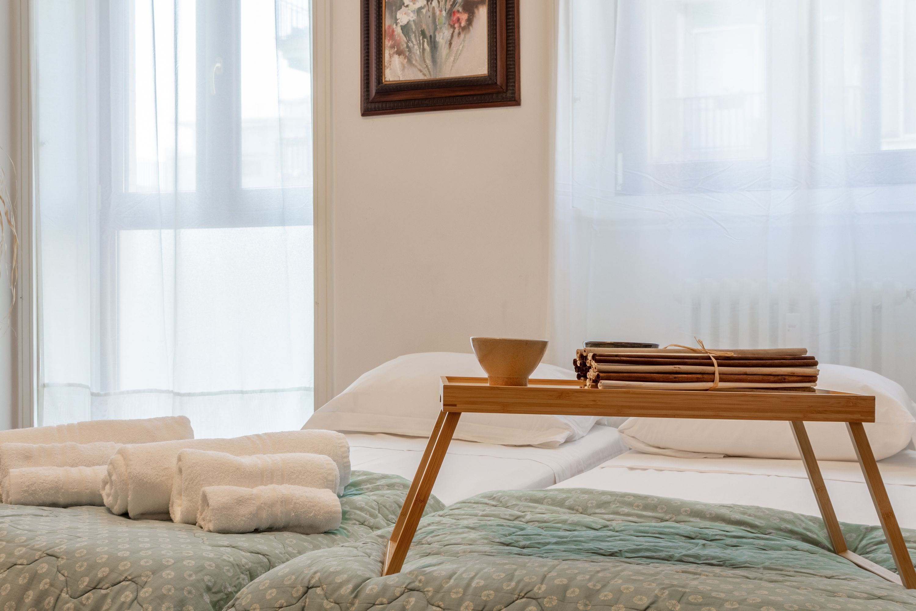 A neatly made bed with a green patterned bedspread, a wooden bedside table with a bowl and rolled-up magazines, and a framed picture on the wall.