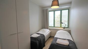 A small, clean room with two single beds, a window with blinds, a desk with a lamp, and a wardrobe.