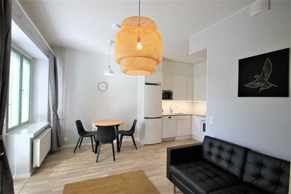 Una sala moderna con un sofá de cuero negro, una mesa de comedor redonda con dos sillas, una lámpara colgante naranja y una cocina al fondo.
