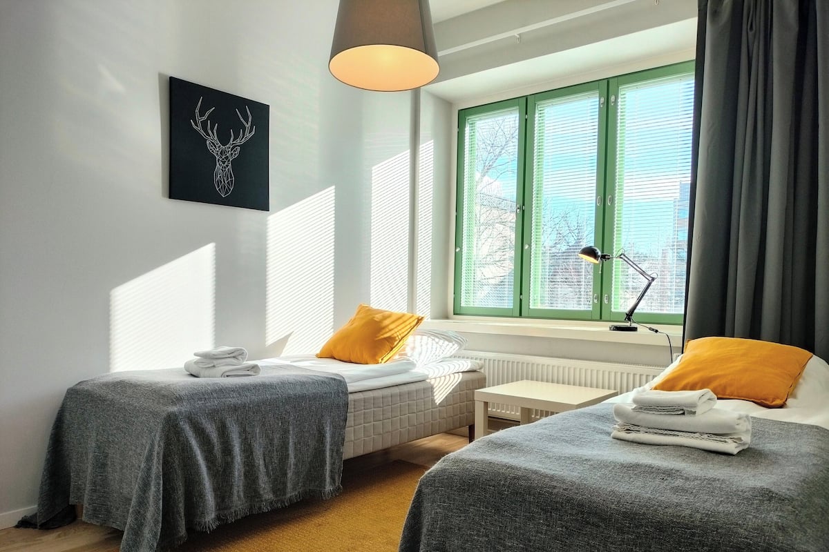 Habitación de hotel con dos camas, una ventana con persianas, un radiador y un cuadro colgado en la pared.