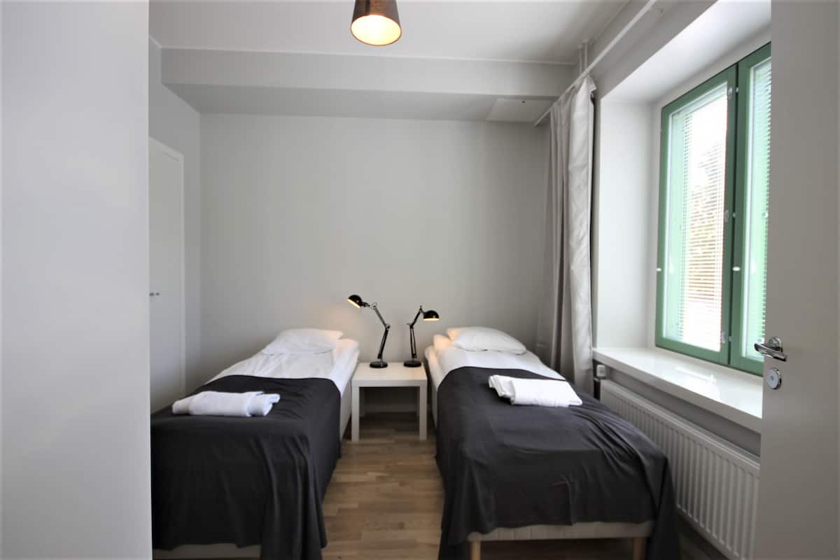 Una habitación con dos camas individuales, una mesita de noche con dos lámparas, un ventanal con contraventanas verdes y un radiador.