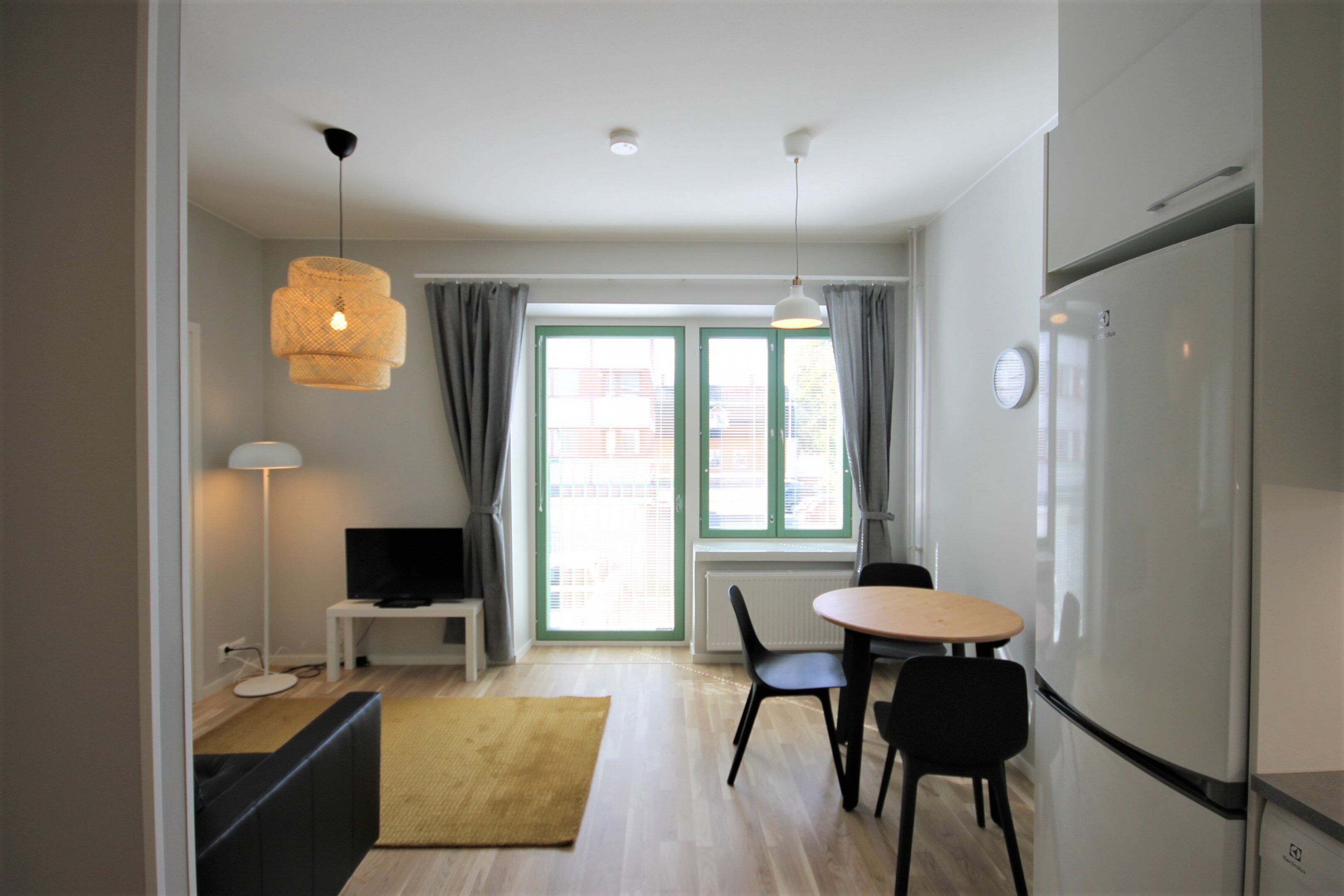 Una cocina moderna con refrigerador, una mesa de comedor con sillas, un televisor sobre un mueble bajo y una ventana con cortinas.