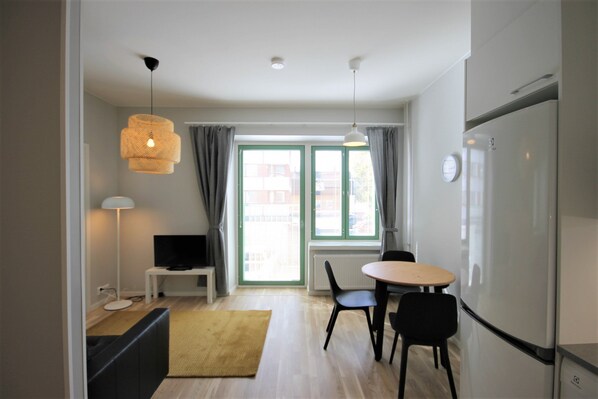Una cocina moderna con refrigerador, una mesa de comedor con sillas, un televisor sobre un mueble bajo y una ventana con cortinas.