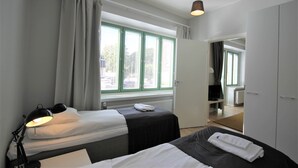 A hotel room with two beds, a desk, a chair, a radiator, and a window with curtains. - Forenom Serviced Apartments Kuopio (Kuopio)