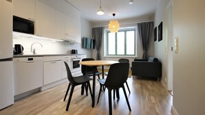 A modern kitchen with white cabinets, a microwave, and a dishwasher. There is a dining area with a round table and black chairs. A large window with curtains is visible, and a sofa is placed near the window. - Forenom Serviced Apartments Kuopio (Kuopio)
