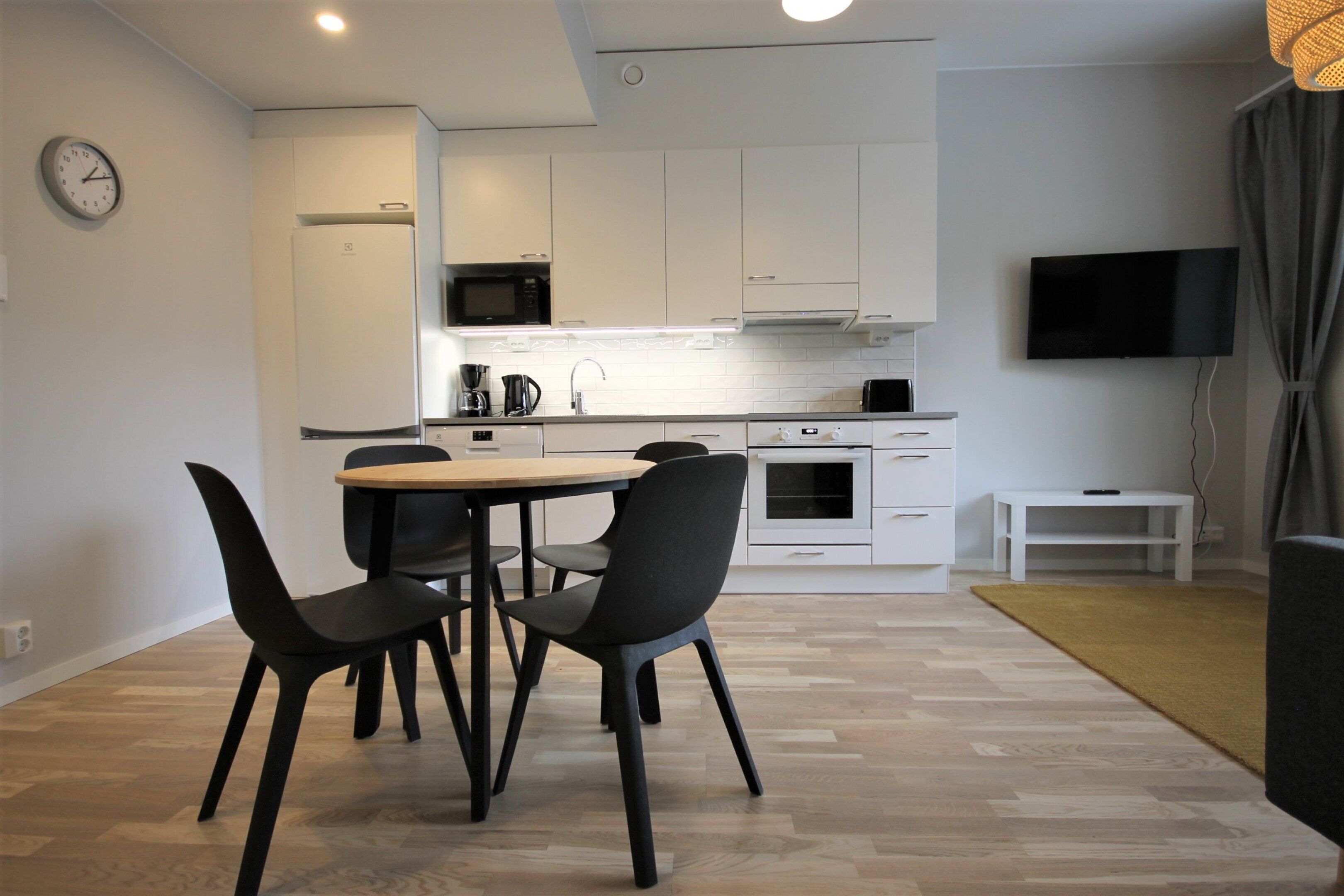 A modern kitchen with white cabinets, a microwave, and a coffee maker. There is a dining table with chairs and a TV mounted on the wall.