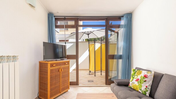 Ein Wohnzimmer mit einer Couch, einem Fernseher auf einem Schrank und einer Glastür, die zu einem Außenbereich mit Sonnenschirm führt.