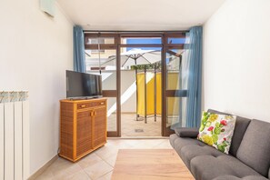 Un salon comprenant un canapé, un téléviseur posé sur un meuble et une porte-fenêtre coulissante donnant sur un espace extérieur agrémenté d’un parasol de terrasse.
