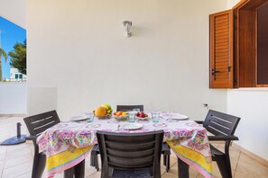 Mesa de jantar posta com pratos, copos e fruta, rodeada por cadeiras, em frente a uma parede branca com persianas de madeira.