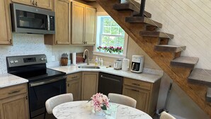 A kitchen with wooden cabinets, a built-in microwave, a stove, a sink, and a coffee maker. There is a round marble table with chairs and a vase of flowers. A wooden staircase is visible in the background. - Lux Cabin 2/2, HotTub, Wood Fireplace, Soaker Tub, Firepit, Walk to Lighthouse (Tremont)