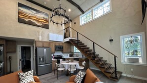 A spacious living area with a modern kitchen, a staircase, and a large window providing natural light. - Lux Cabin 2/2, HotTub, Wood Fireplace, Soaker Tub, Firepit, Walk to Lighthouse (Tremont)