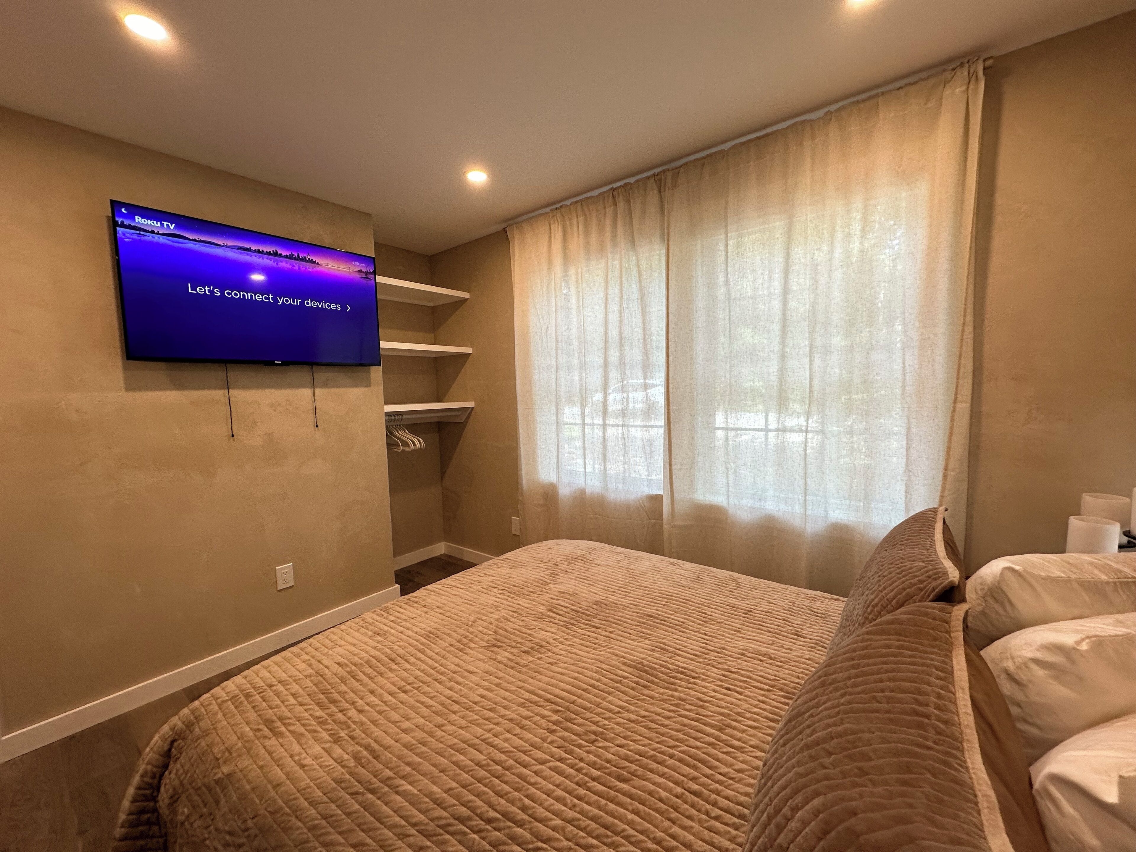 A hotel room with a bed, a TV mounted on the wall, a window with curtains, and a built-in closet with shelves.