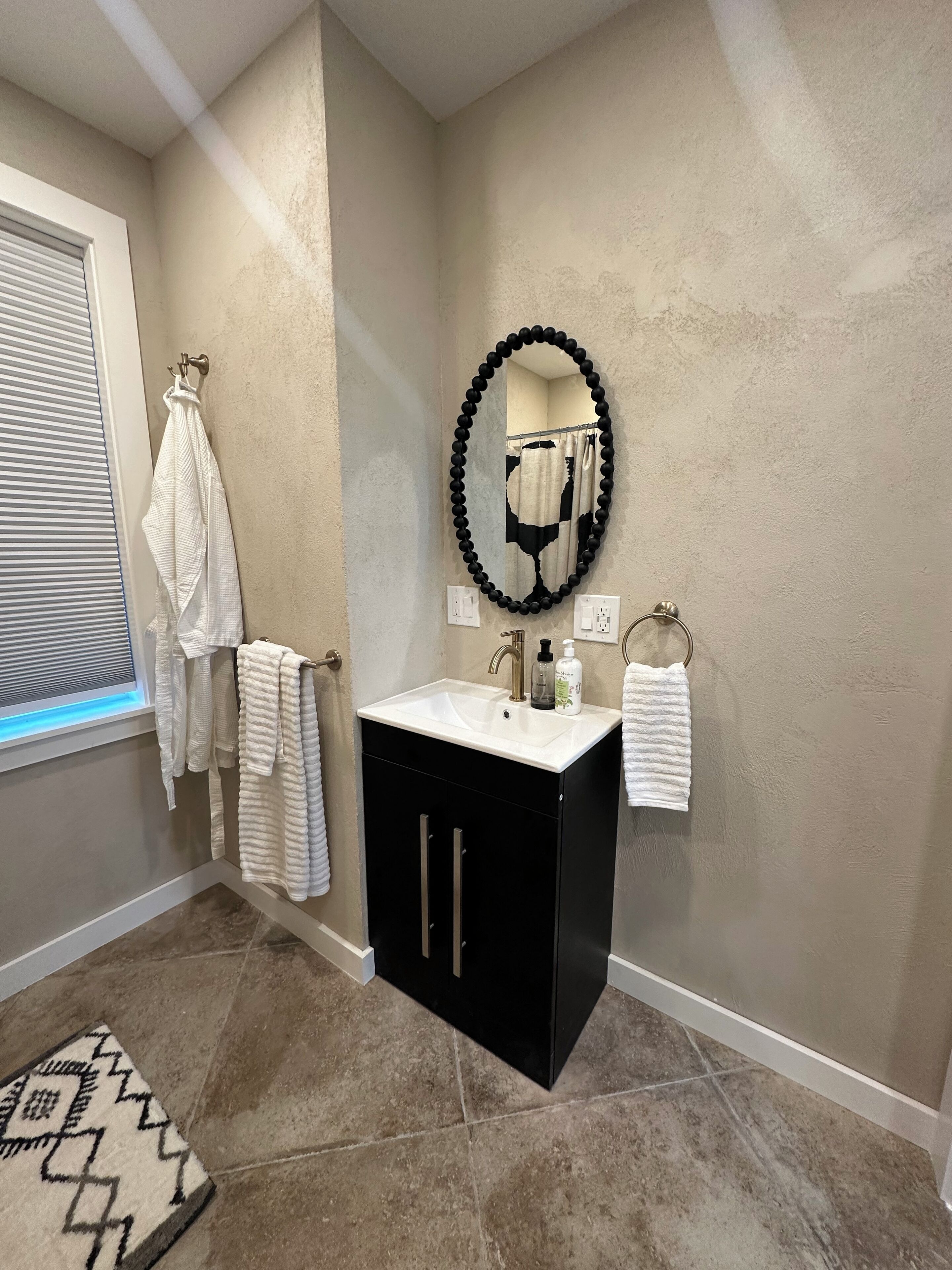 Une salle de bain avec un meuble-lavabo noir, un lavabo blanc et un miroir à cadre noir. Des serviettes sont suspendues à des crochets et un tapis est posé sur le sol.