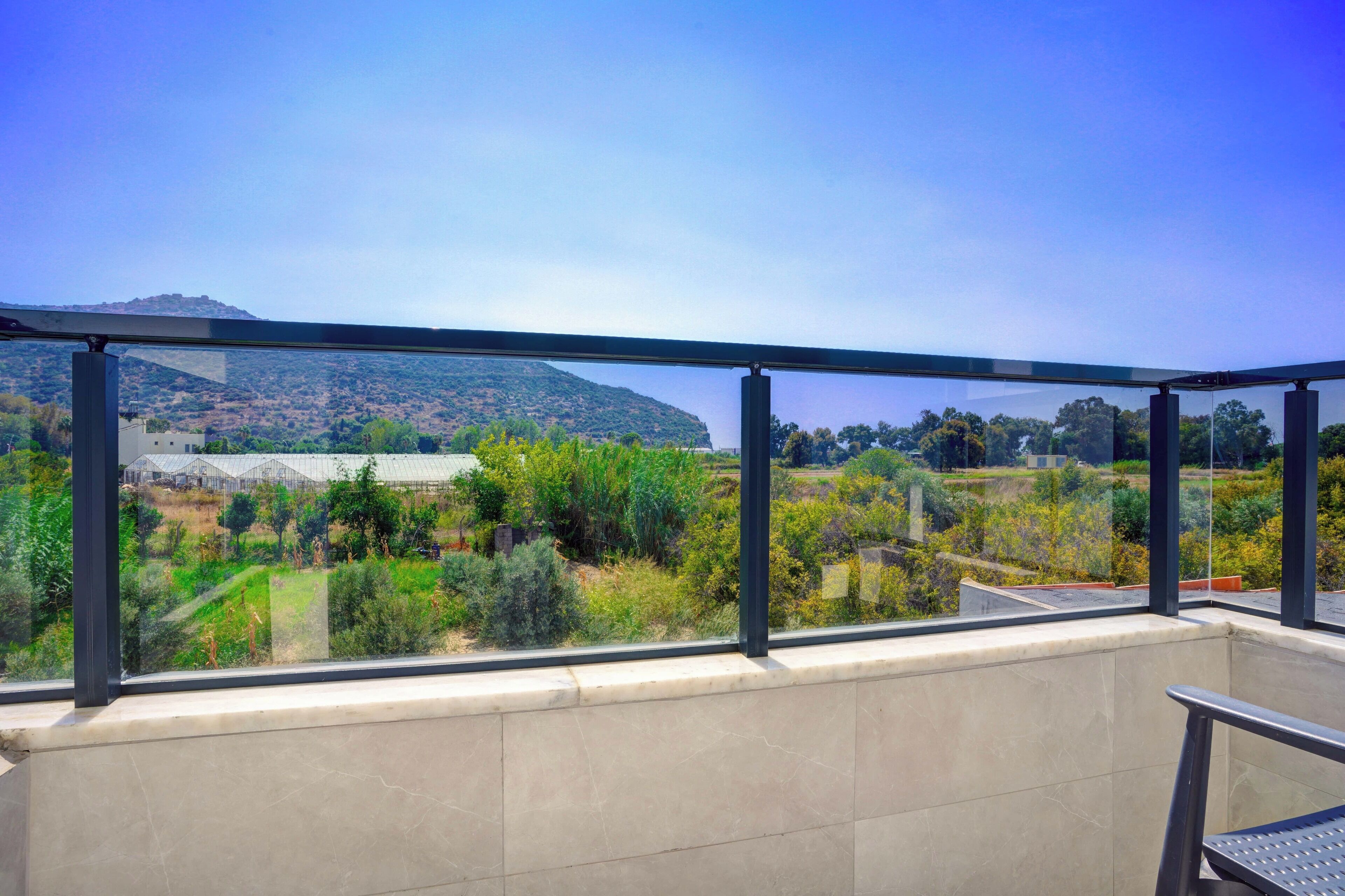 Un balcon doté d’une balustrade en verre, offrant une vue sur un paysage verdoyant et une colline lointaine.