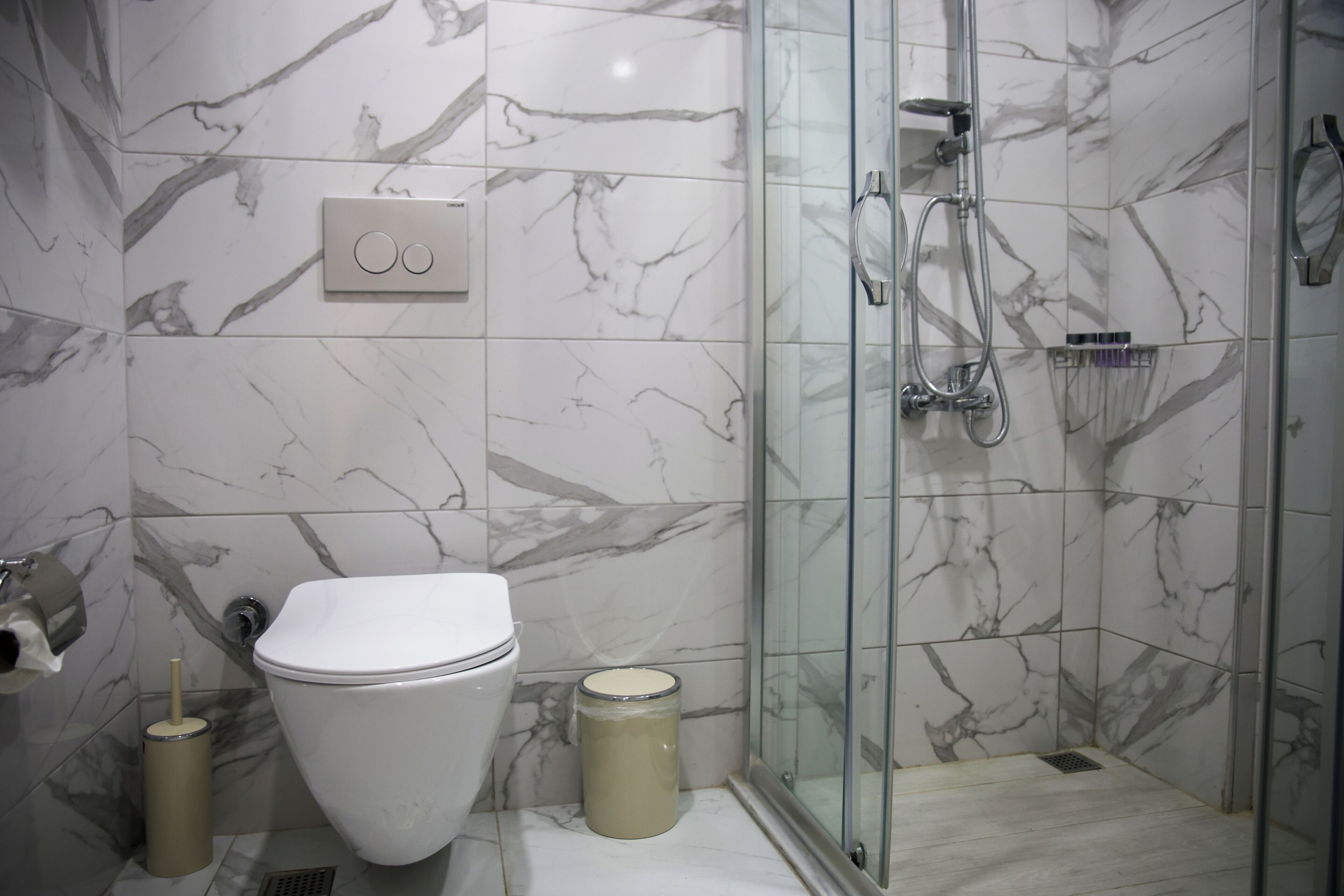 A bathroom with a toilet, a glass shower enclosure, marble tiles, and a trash can.