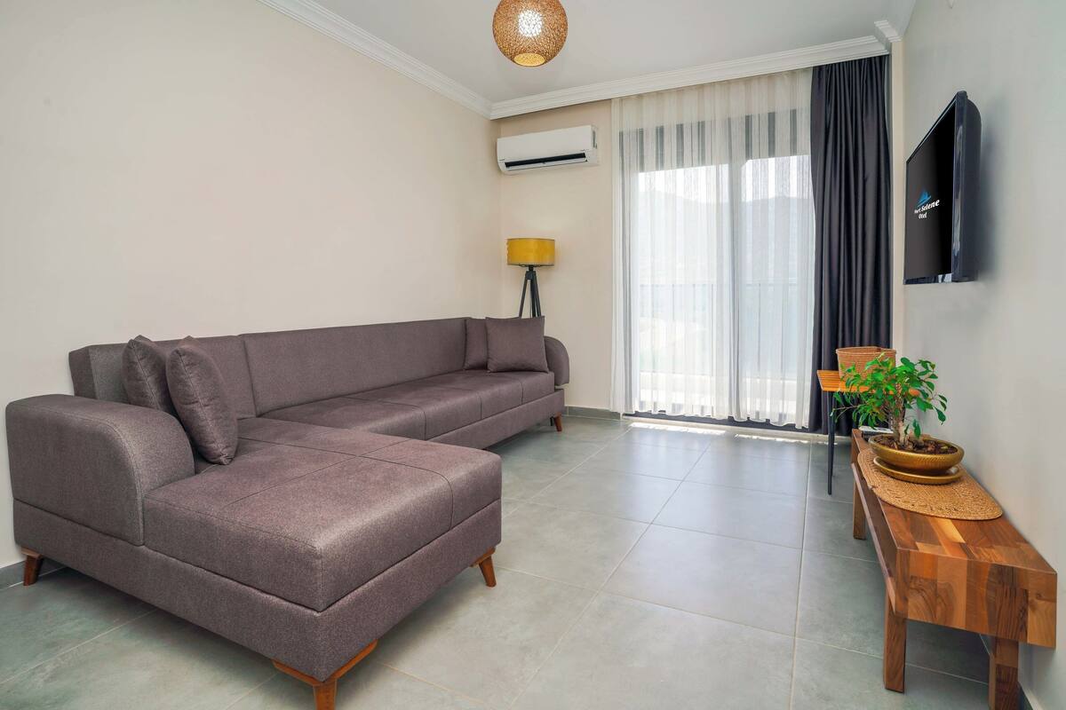 Un salon moderne avec un canapé d’angle, un banc en bois, une plante en pot, une lampe sur pied et un téléviseur fixé au mur.