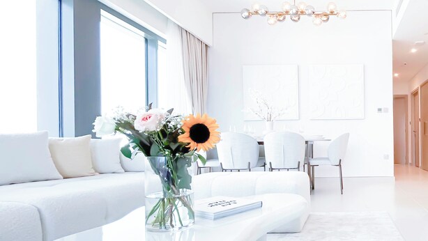 Een moderne woonkamer met een witte bank, een glazen salontafel en een vaas met bloemen. Er is een eethoek met witte stoelen en een kroonluchter.