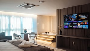 Habitación de hotel moderna con una gran televisión de pantalla plana mostrando una guía de canales, una cama con toallas ordenadas, un área de descanso con sillas y una escalera que lleva a un balcón.