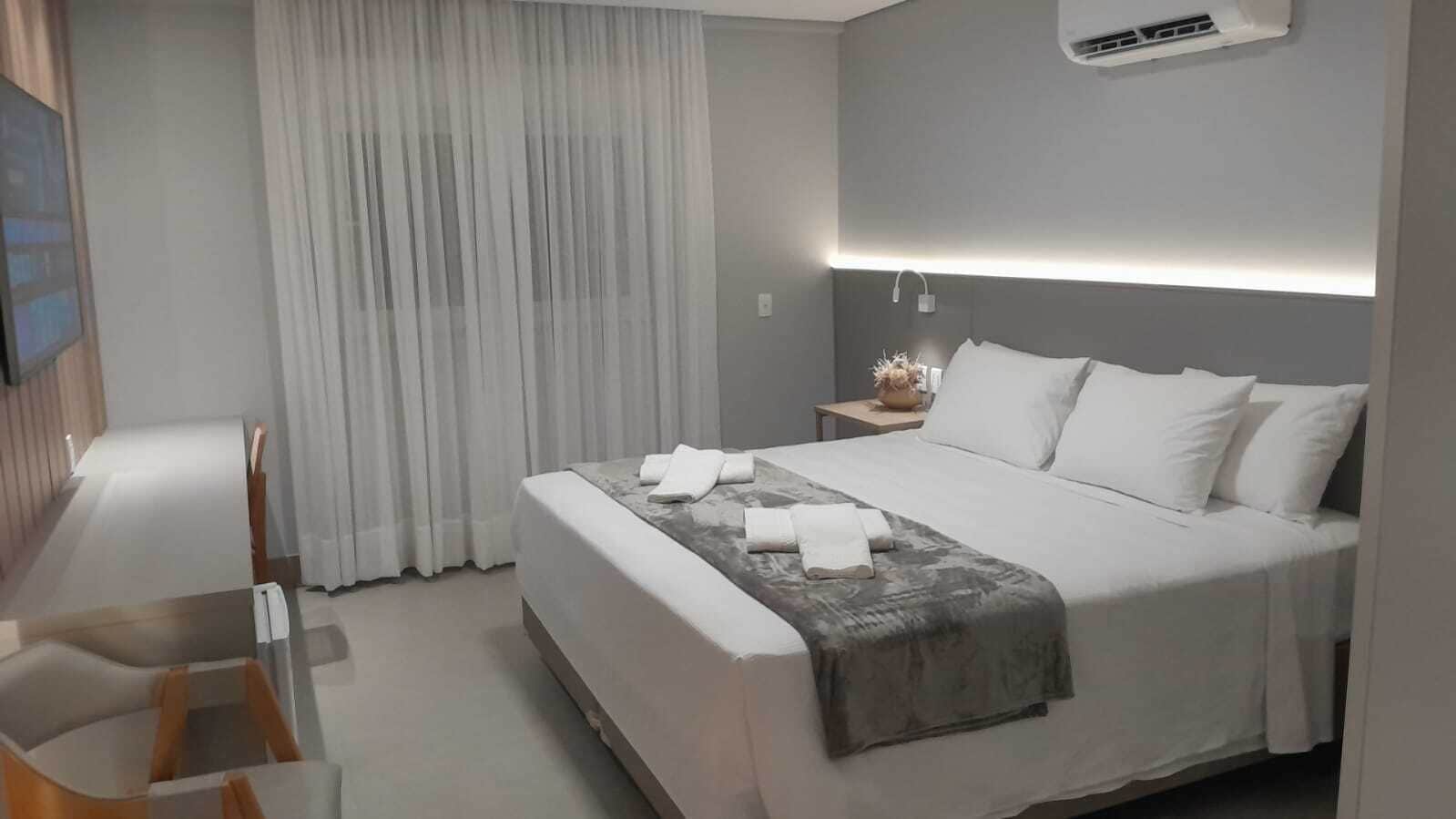 Ein modernes Hotelzimmer mit einem ordentlich bezogenen Bett, einem Nachttisch mit Lampe und einem großen Fenster mit Vorhängen.