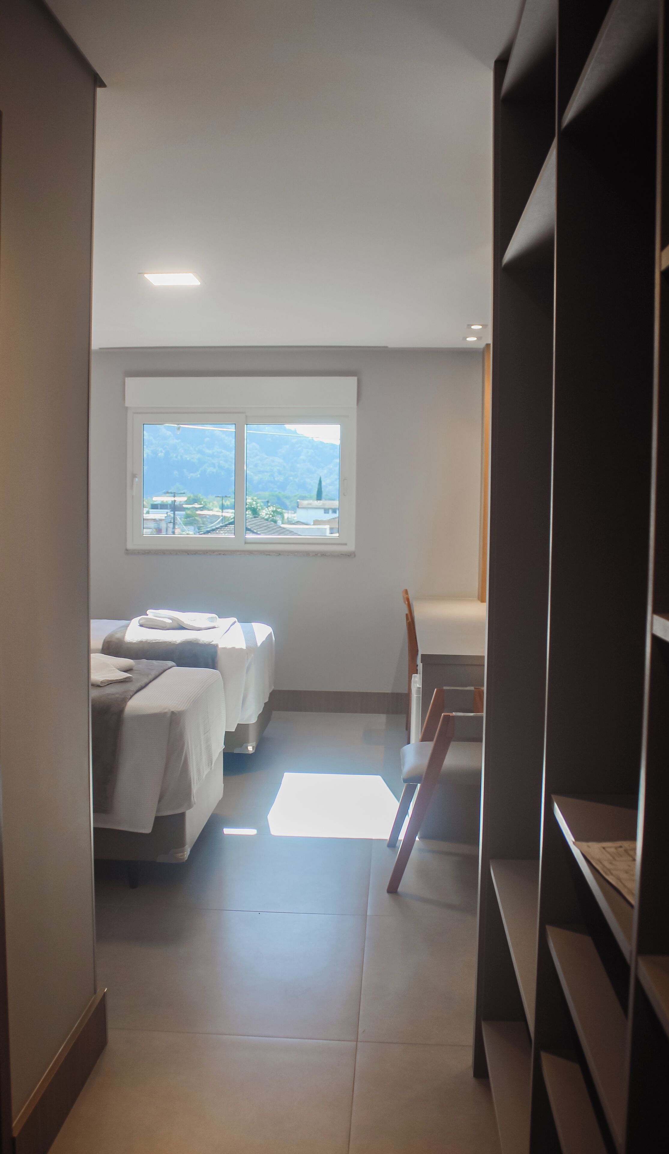 Un couloir étroit avec un lit, un bureau et une fenêtre donnant sur des immeubles et des montagnes.
